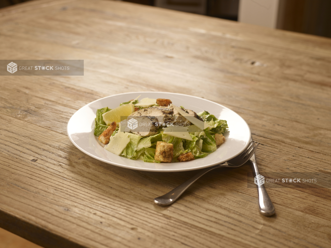 Grilled Chicken Caesar Salad with Shaved Parmesan Cheese, Croutons and a Lemon Wedge on a Round White Ceramic Dish on a Rustic Wooden Surface