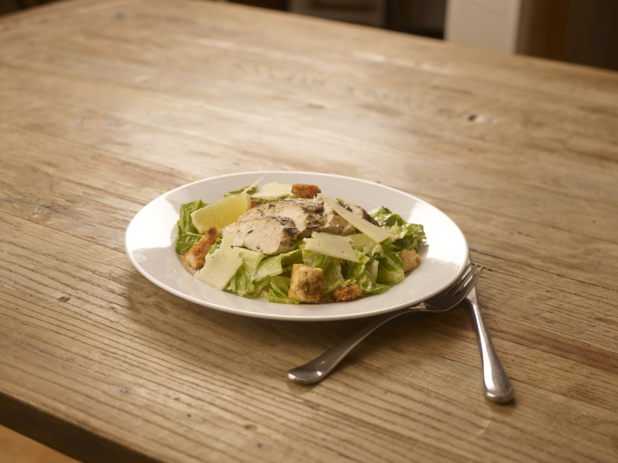 Grilled Chicken Caesar Salad with Shaved Parmesan Cheese, Croutons and a Lemon Wedge on a Round White Ceramic Dish on a Rustic Wooden Surface