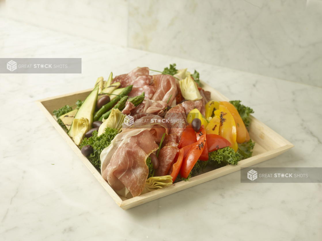 Antipasto Tray Featuring Italian Cold Cuts, Roasted Peppers, Grilled Vegetables and Artichoke Hearts on a Bed of Kale in a Square Wood Serving Tray on a Marble Surface