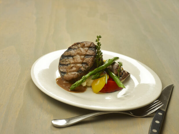 Grilled Ribeye Steak Medallion with Au Jus and a Side of Colourful Cooked Vegetables (Asparagus, Red and Yellow Peppers, Portobello Mushroom, Cauliflower) on a White Ceramic Dish on a Wooden Surface