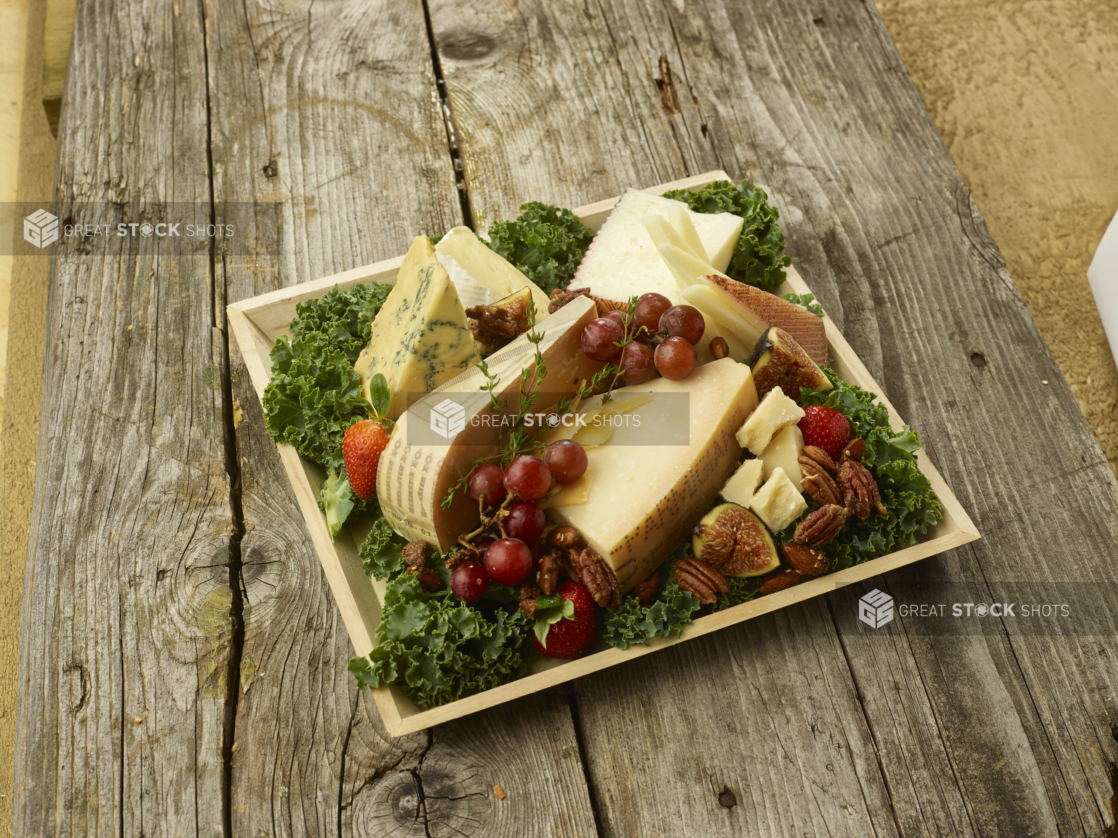 Assorted Sliced and Cut Cheeses with an Assortment of Fruit and Nuts on a bed of Kale Leaves in a Square Wooden Tray on an Aged Wooden Table