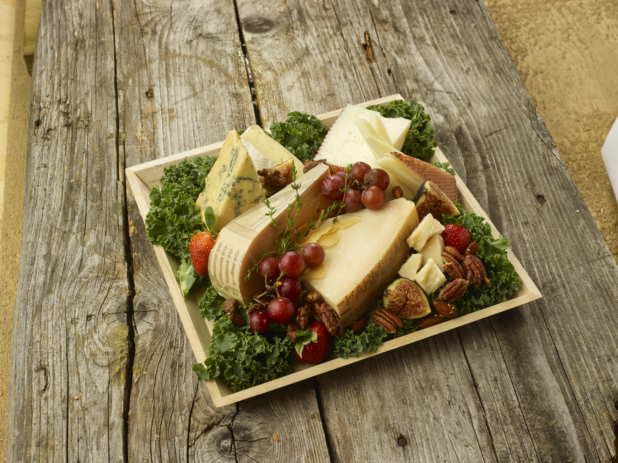 Assorted Sliced and Cut Cheeses with an Assortment of Fruit and Nuts on a bed of Kale Leaves in a Square Wooden Tray on an Aged Wooden Table