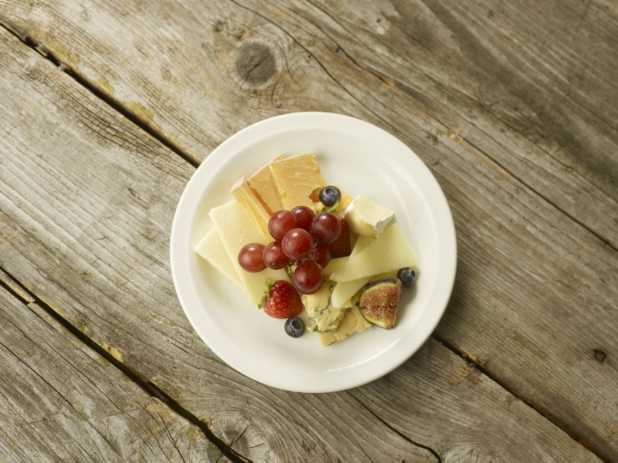 Overhead View of Assorted Sliced Cheeses (Blue, Brie, Parmesan, Swiss, Cheddar) and Fresh Fruits on a Round White Ceramic Dish on a Rustic Wooden Surface