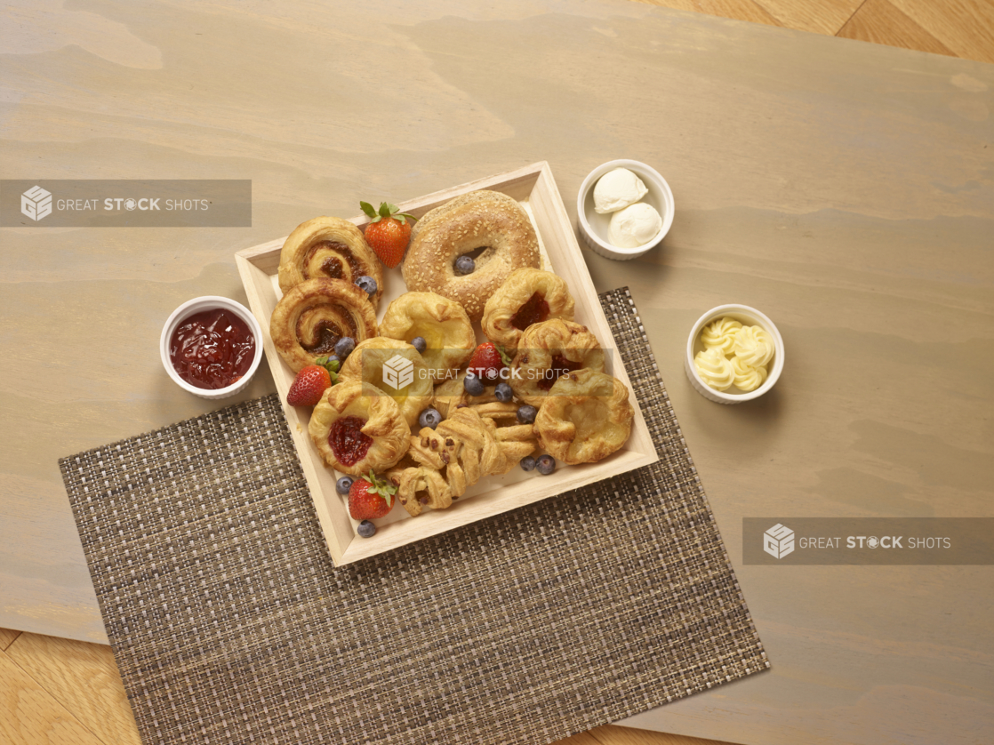 Overhead View of Square Wooden Tray with Assorted Danishes, Cinnamon Rolls and Bagels on a Wooden Table with Jam and Butter – for Breakfast Catering