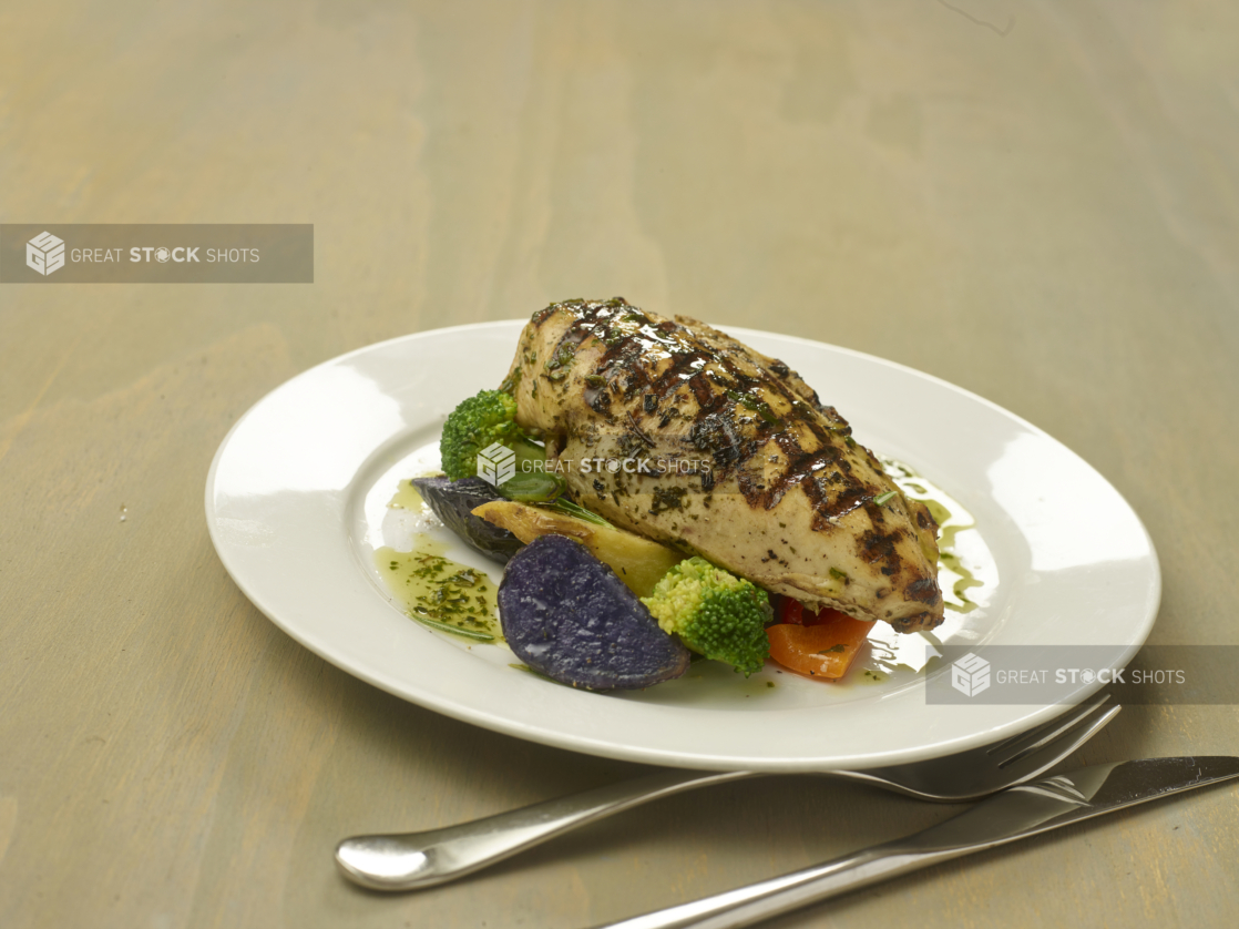 Lemon Herbed Grilled Chicken Breast with Cooked Vegetables (Broccoli, Potatoes, Peppers) on a White Ceramic Dish on a Wooden Surface