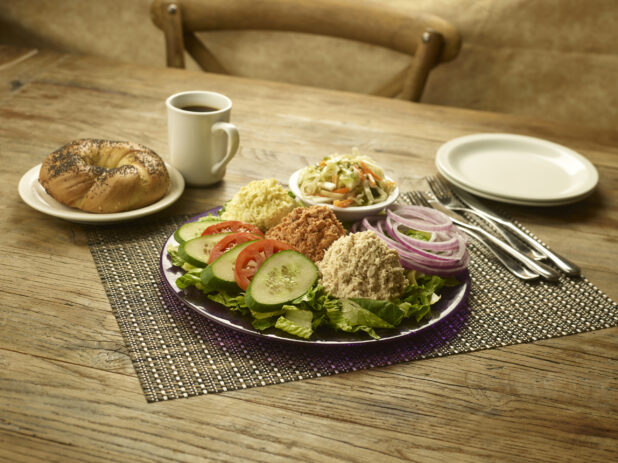 Dairy platter, egg salad, tuna salad, salmon salad in mounds on a plate with a side of coleslaw, sliced red onion, tomato and cucumber and a whole bagel and a cup of coffee in the background