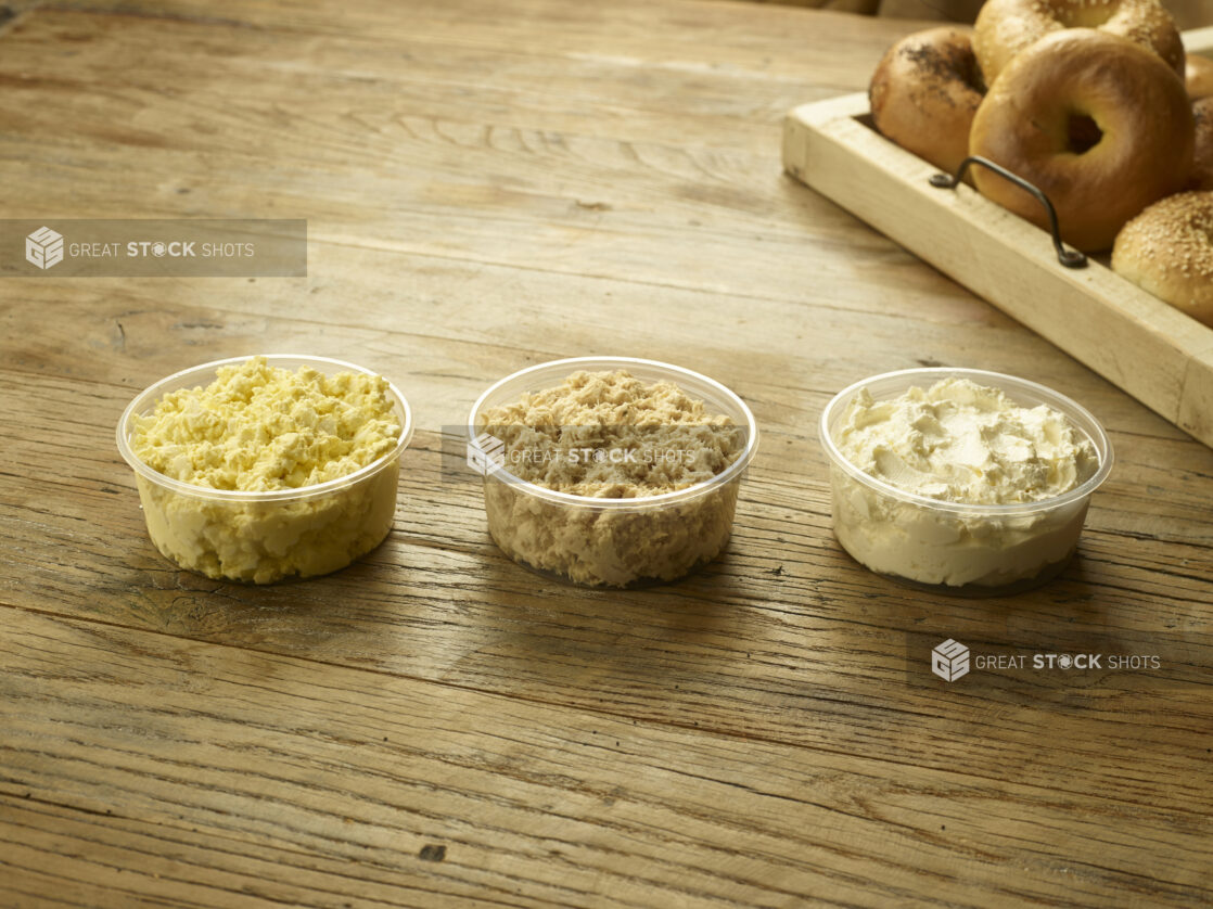 3x250 ml clear plastic containers with egg salad, tuna salad and cream cheese with a wooden tray of assorted bagels in the background all on a rustic wooden table