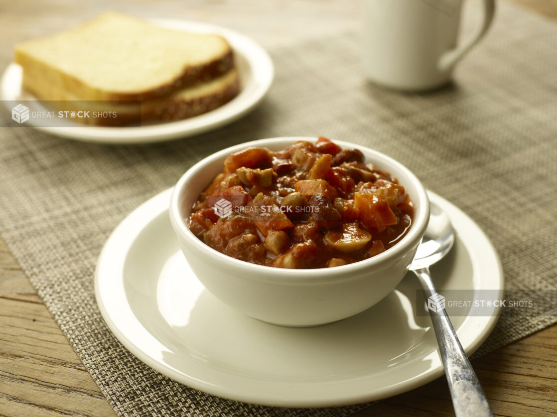 Vegetarian chili in a small white bowl on a white side plate with a spoon, bread and a coffee cup in the background