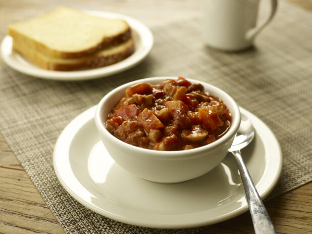 Vegetarian chili in a small white bowl on a white side plate with a spoon, bread and a coffee cup in the background
