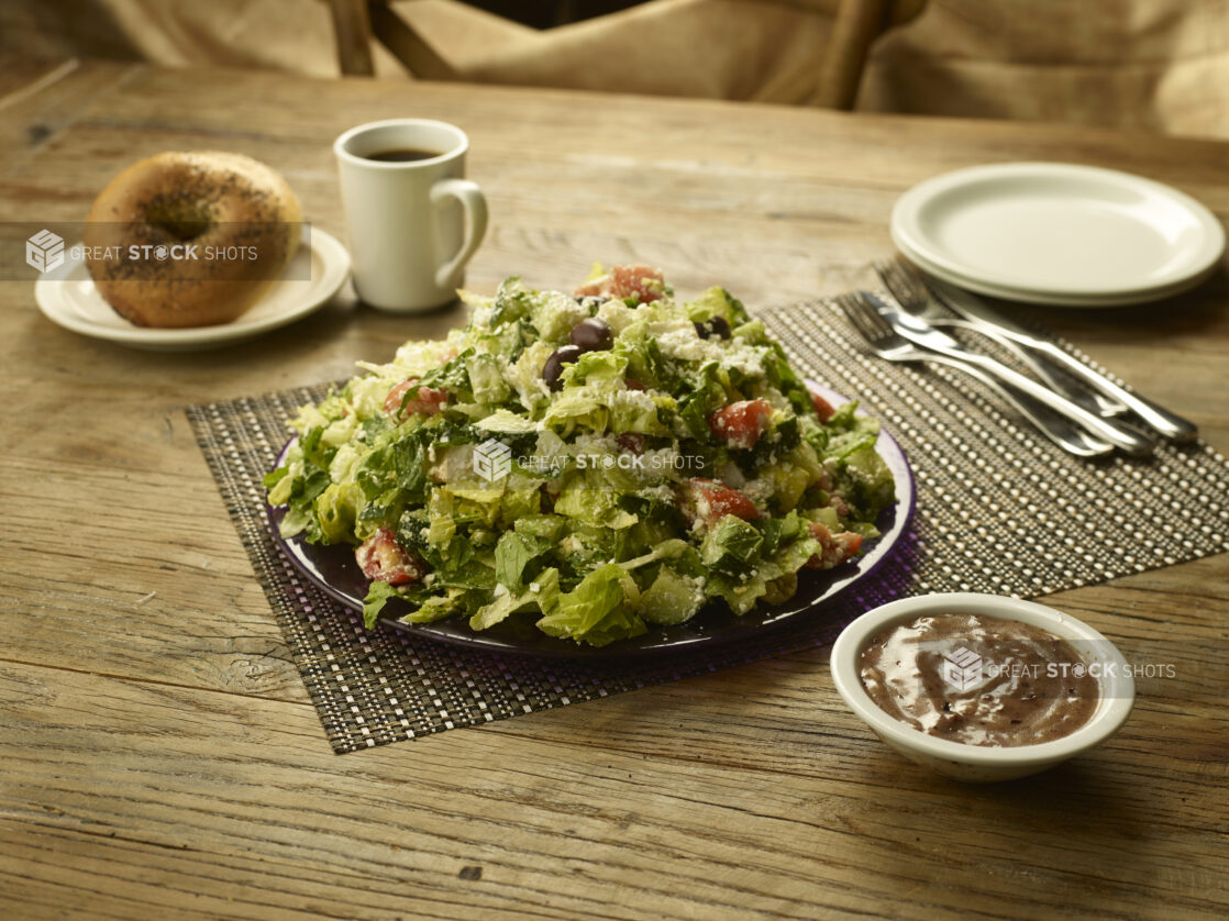 Greek salad on a place setting with dressing in a ramekin in the foreground and bagels and a cup of coffee in the background with white side plates and cutlery