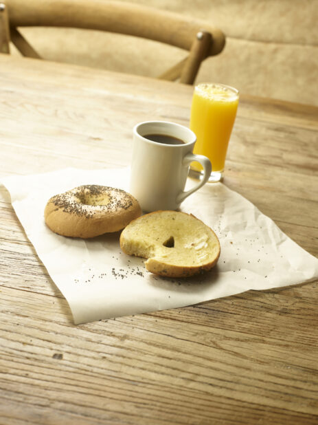 A toasted bagel sliced in half with butter on parchment paper with a cup of coffee and a glass of orange juice in the background  all on a rustic wooden table