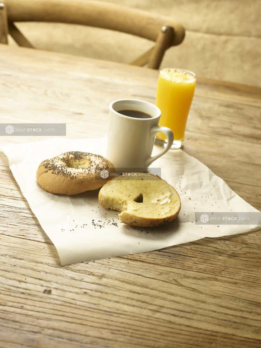 A toasted bagel sliced in half with butter on parchment paper with a cup of coffee and a glass of orange juice in the background  all on a rustic wooden table