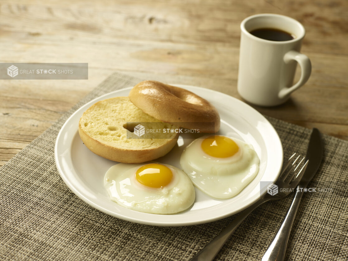 2 eggs sunny side up with a toasted bagel on a white round plate on a placemat with a knife and fork and a cup of coffee in the background