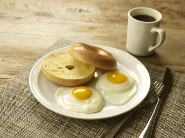 2 eggs sunny side up with a toasted bagel on a white round plate on a placemat with a knife and fork and a cup of coffee in the background