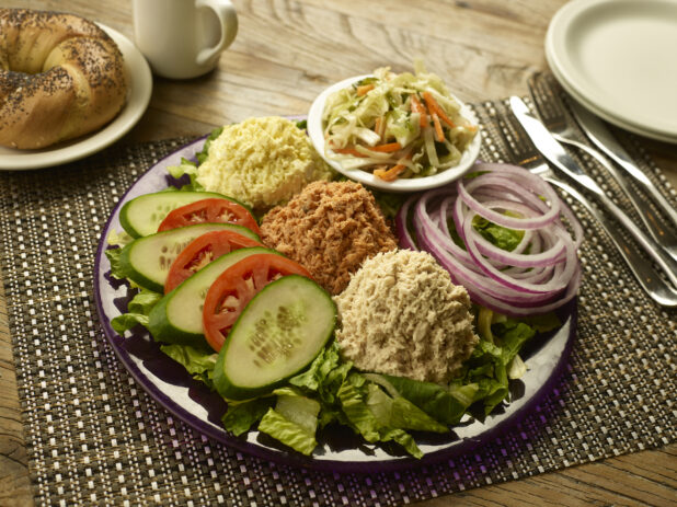 Dairy platter, egg salad, tuna salad, salmon salad in mounds on a plate with a side of coleslaw, sliced red onion, tomato and cucumber and a whole bagel and a cup of coffee in the background