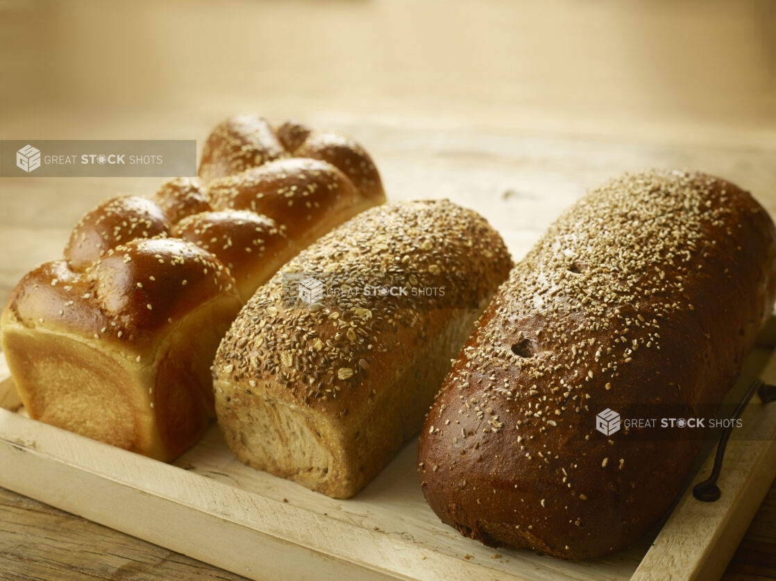 3 whole loaves of fresh bread (challah, multigrain and pumpernickel) on a wooden tray, on a wooden background