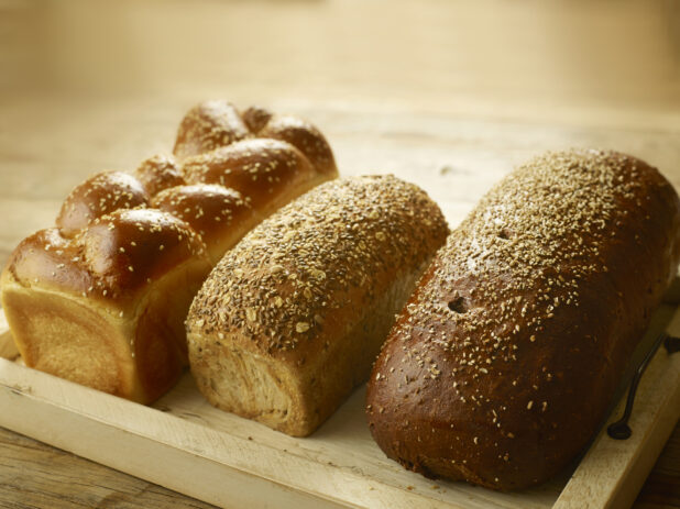 3 whole loaves of fresh bread (challah, multigrain and pumpernickel) on a wooden tray, on a wooden background