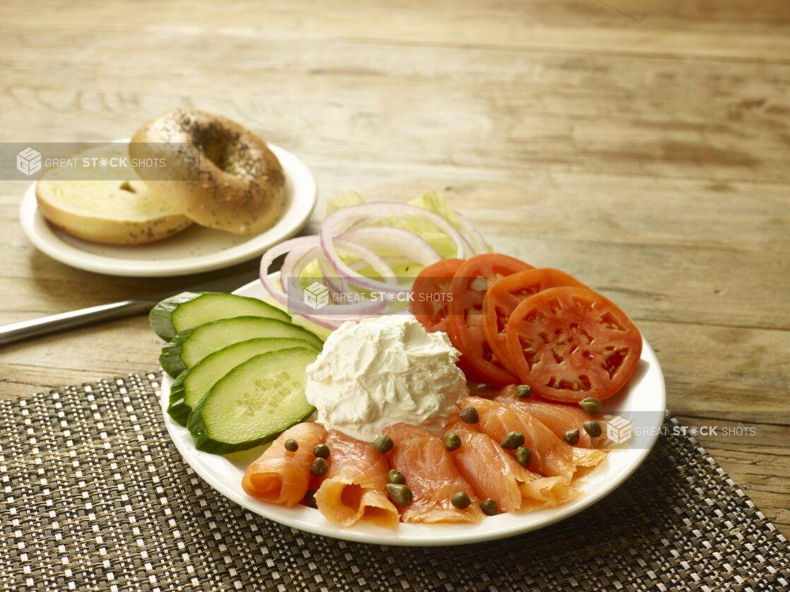 Smoked salmon with capers on a round white plate with sliced tomatoes, cucumbers, onions and lettuce with a scoop of cream cheese and a toasted bagel on the side