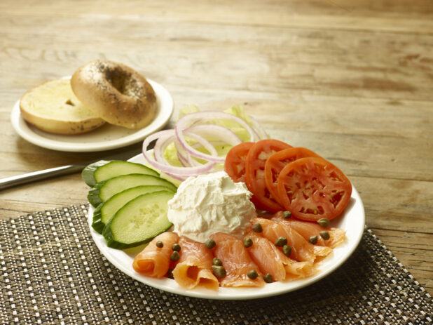 Smoked salmon with capers on a round white plate with sliced tomatoes, cucumbers, onions and lettuce with a scoop of cream cheese and a toasted bagel on the side