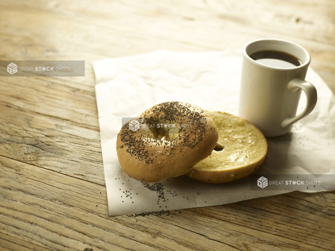 A toasted bagel sliced in half with butter on parchment paper with a cup of coffee in the background  all on a rustic wooden table