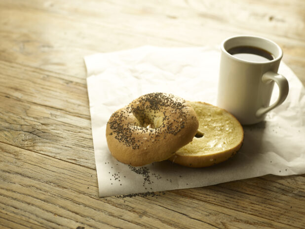 A toasted bagel sliced in half with butter on parchment paper with a cup of coffee in the background  all on a rustic wooden table