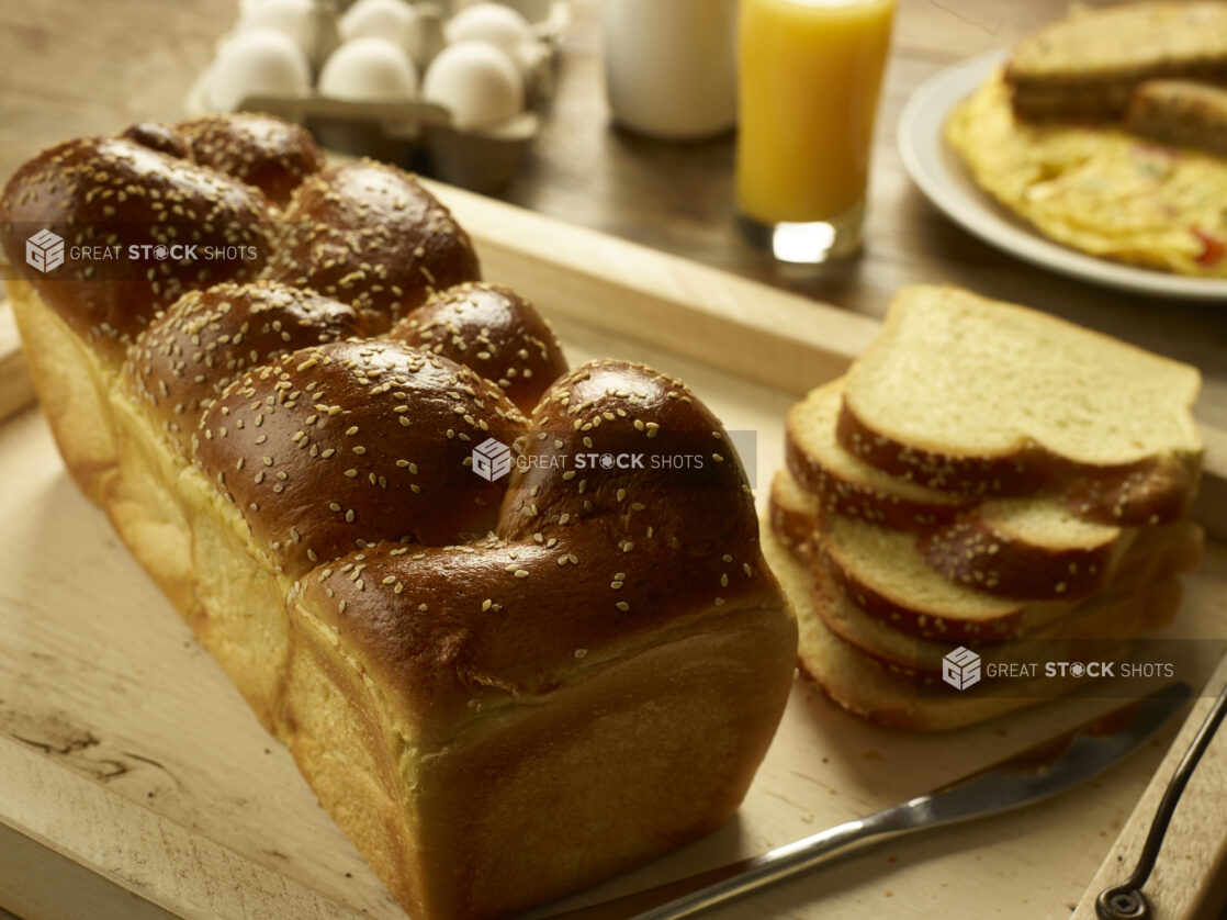 Whole and sliced challah, egg bread, on a wooden tray in the foreground with a carton of eggs, a glass of orange juice and an omelet and toast in the background