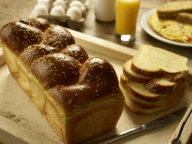 Whole and sliced challah, egg bread, on a wooden tray in the foreground with a carton of eggs, a glass of orange juice and an omelet and toast in the background
