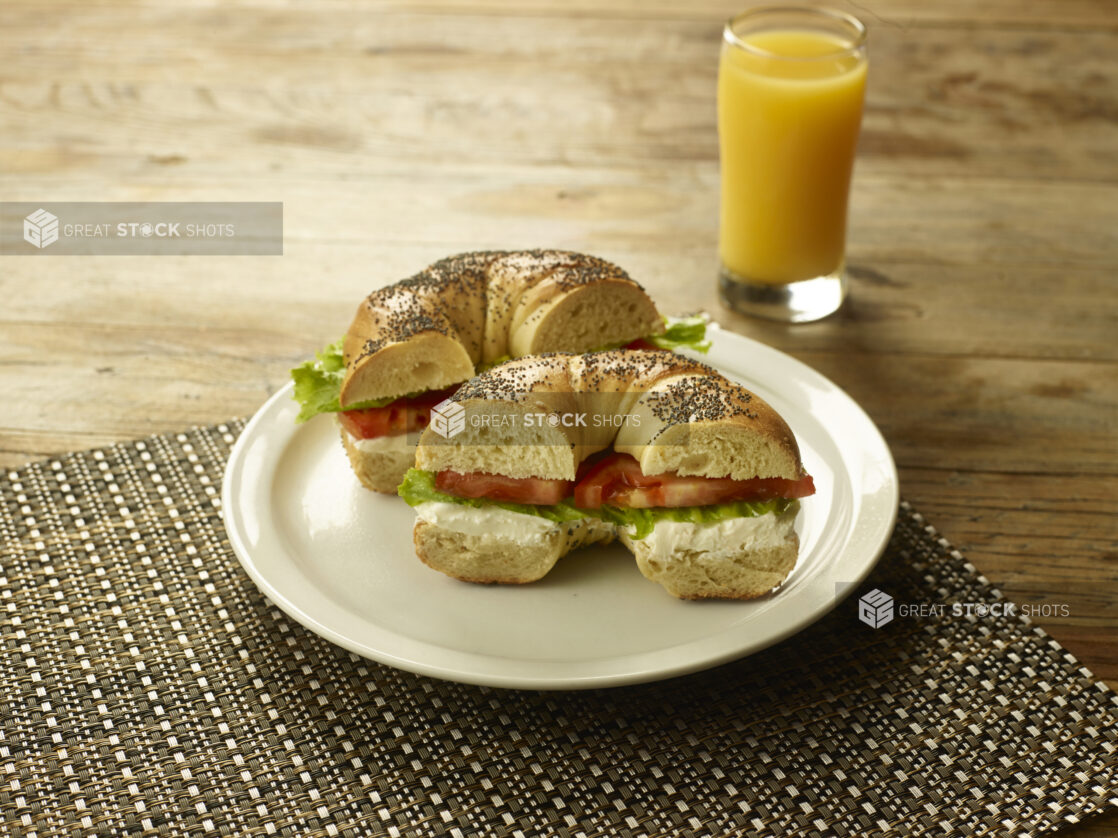 Cream Cheese, Lettuce and Tomato Sandwich on a Poppy Seed Bagel on a Round White Ceramic Dish on a Woven Placemat with a Glass of Orange Juice on a Rustic Wooden Table