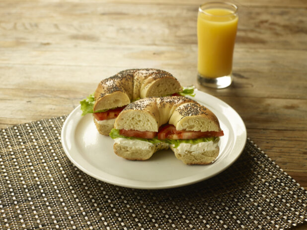 Cream Cheese, Lettuce and Tomato Sandwich on a Poppy Seed Bagel on a Round White Ceramic Dish on a Woven Placemat with a Glass of Orange Juice on a Rustic Wooden Table