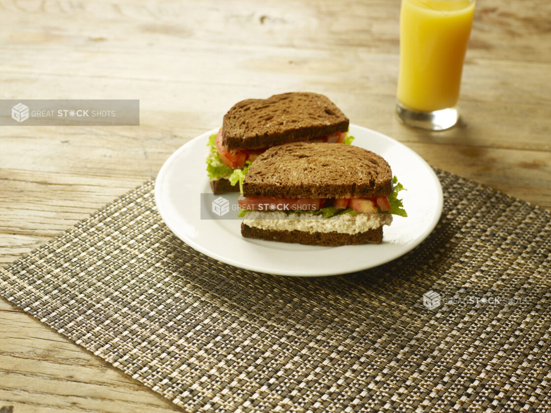 Tuna Salad Sandwich with Lettuce and Tomato on Dark Rye Bread on a Round White Ceramic Dish on a Woven Placemat with a Glass of Orange Juice on a Rustic Wooden Table