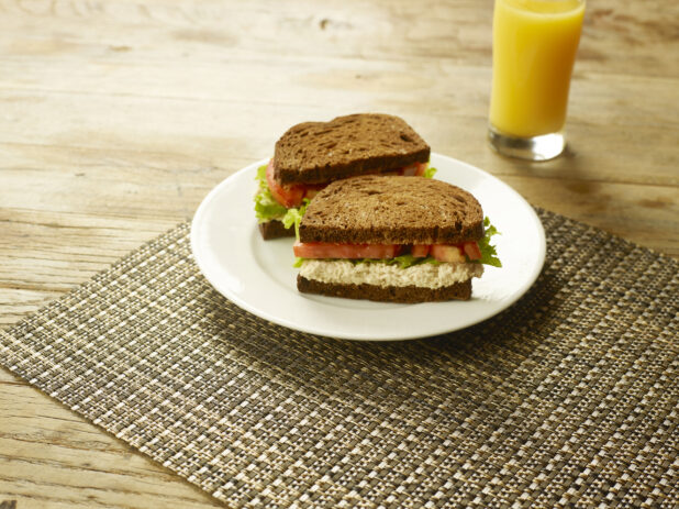 Tuna Salad Sandwich with Lettuce and Tomato on Dark Rye Bread on a Round White Ceramic Dish on a Woven Placemat with a Glass of Orange Juice on a Rustic Wooden Table