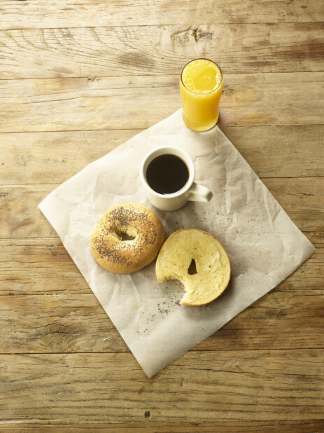 Overhead view of a toasted bagel sliced in half with butter on parchment paper with a cup of coffee and a glass of orange juice in the background  all on a rustic wooden table