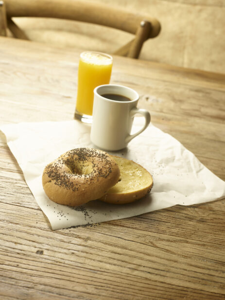 A toasted bagel sliced in half with butter on parchment paper with a cup of coffee and a glass of orange juice in the background  all on a rustic wooden table