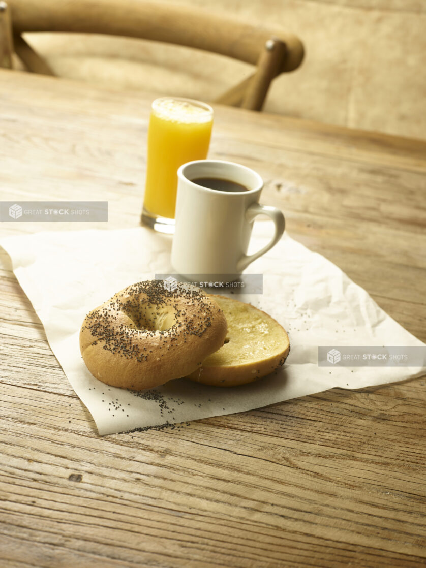 A toasted bagel sliced in half with butter on parchment paper with a cup of coffee and a glass of orange juice in the background  all on a rustic wooden table