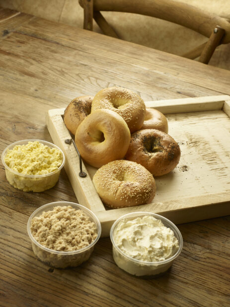 A Spread of Assorted Bagels and Dairy Platter Items - Egg Salad, Tuna Salad and Cream Cheese - on a Wooden Tray on a Rustic Wooden Table in an Indoor Setting