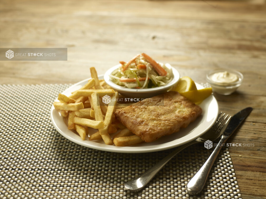 Fish and chips meal with coleslaw and a side of tartar sauce on a placemat with cutlery on a wooden table