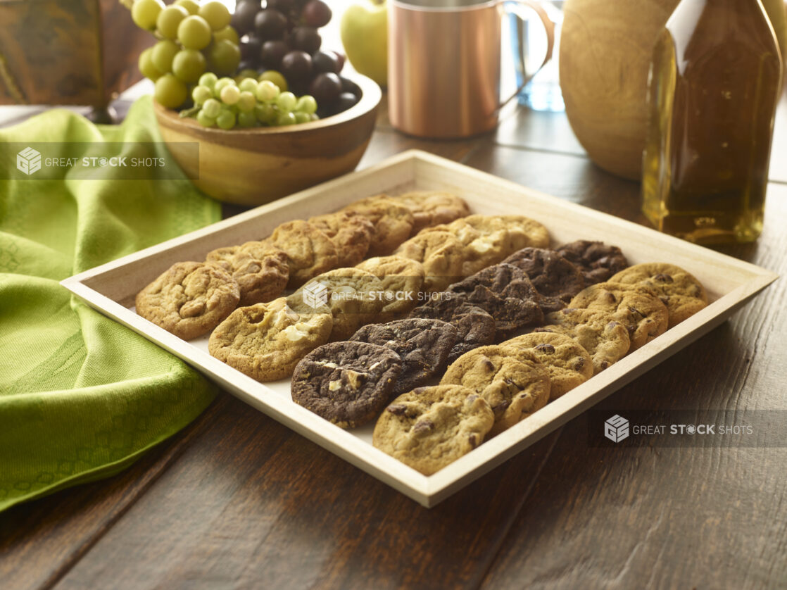 Assorted cookie tray on a wooden catering tray with a wooden bowl of grapes and other accessories in the background on a rustic wooden table