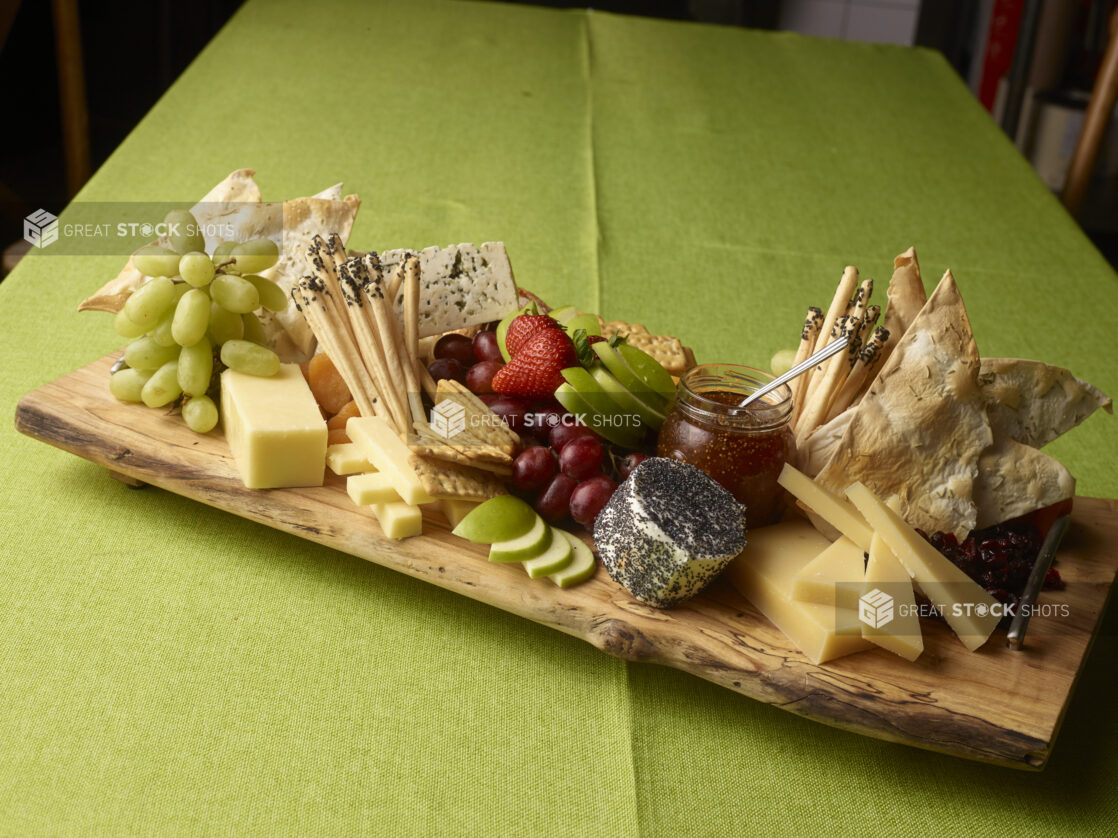 Elegant cheese tray with various cheeses and crackers with fresh fruit and fig jam in a jar on a wooden serving tray on a lime green tablecloth