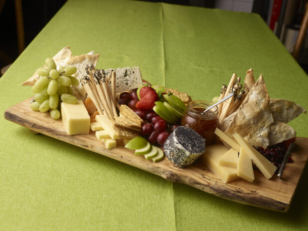 Elegant cheese tray with various cheeses and crackers with fresh fruit and fig jam in a jar on a wooden serving tray on a lime green tablecloth