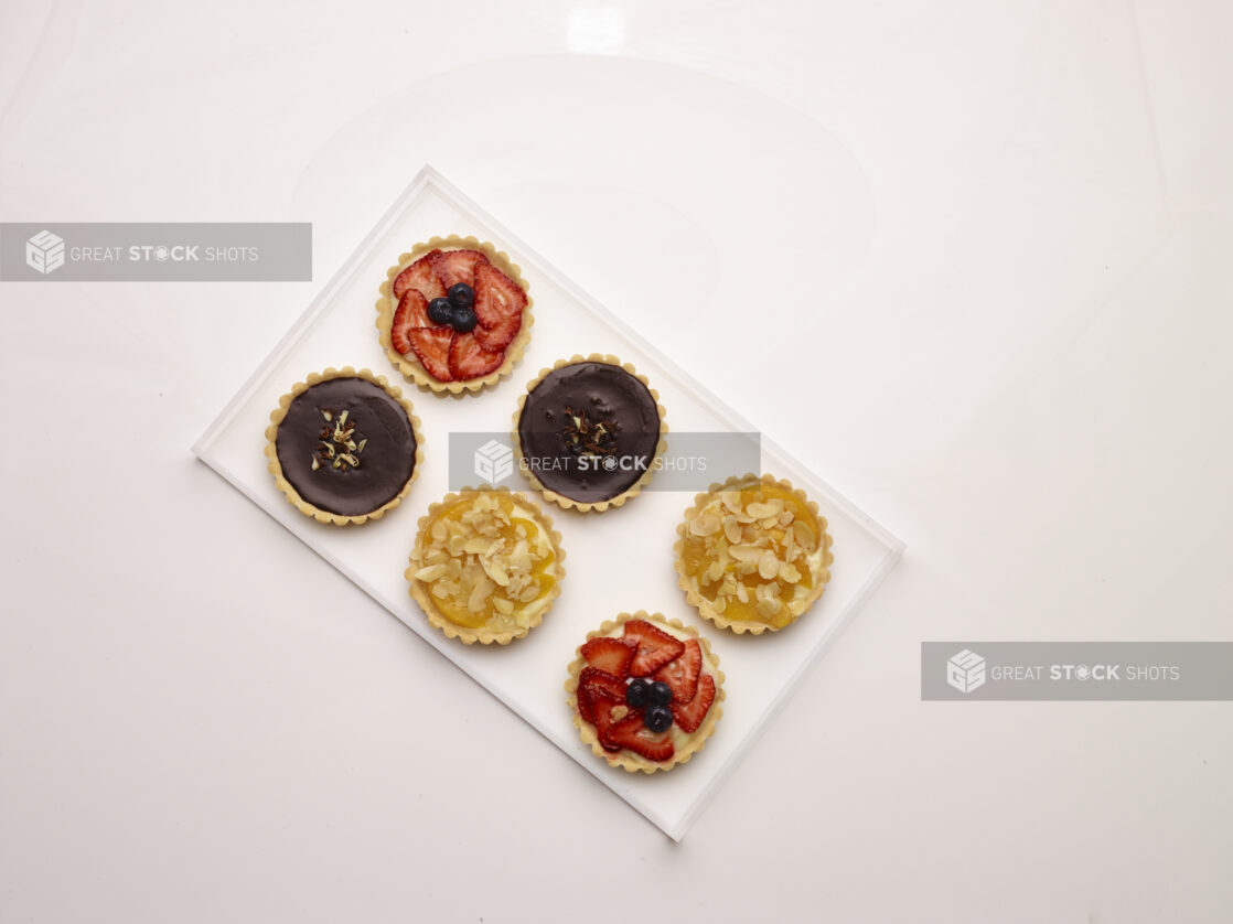 Overhead view of assorted sweet tarts, chocolate, strawberry flan and peach with slivered almonds, on a white tray on a white background