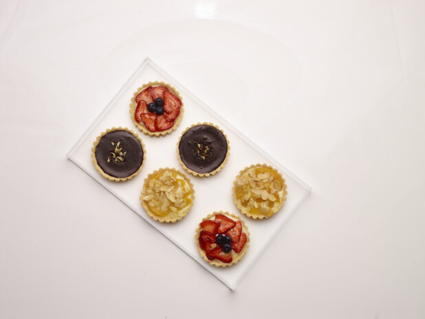 Overhead view of assorted sweet tarts, chocolate, strawberry flan and peach with slivered almonds, on a white tray on a white background