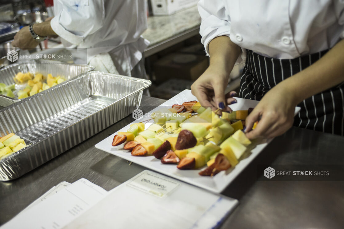 Chef's Hands Preparing a Large order of Fresh Fruit Skewers on a Square White Ceramic Plate in a Kitchen Setting of a Restaurant, for Catering or a Party