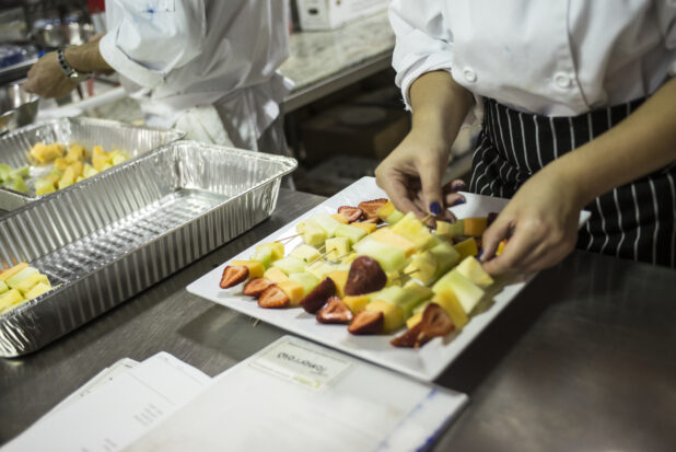 Chef's Hands Preparing a Large order of Fresh Fruit Skewers on a Square White Ceramic Plate in a Kitchen Setting of a Restaurant, for Catering or a Party