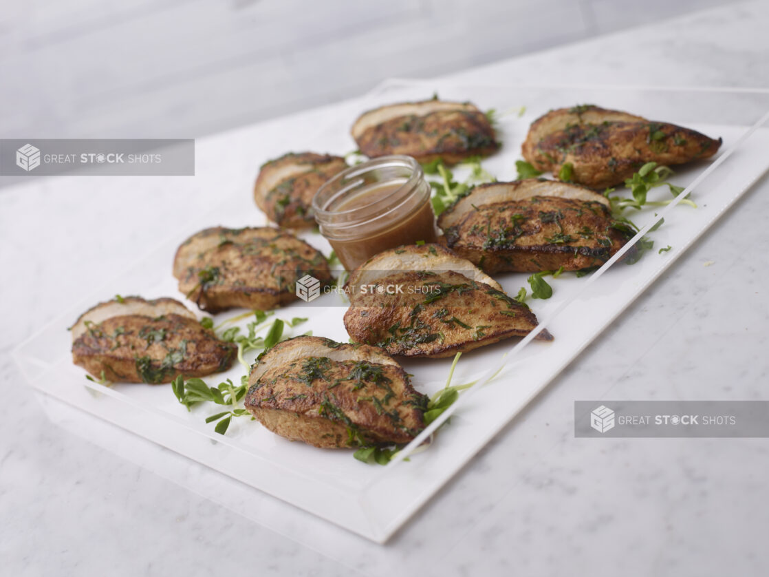 Sliced Herb-Roasted Pork Medallions with a Jar of Au Jus Sauce in a Clear Plexiglass Platter on a Marble Surface in an Indoor Setting