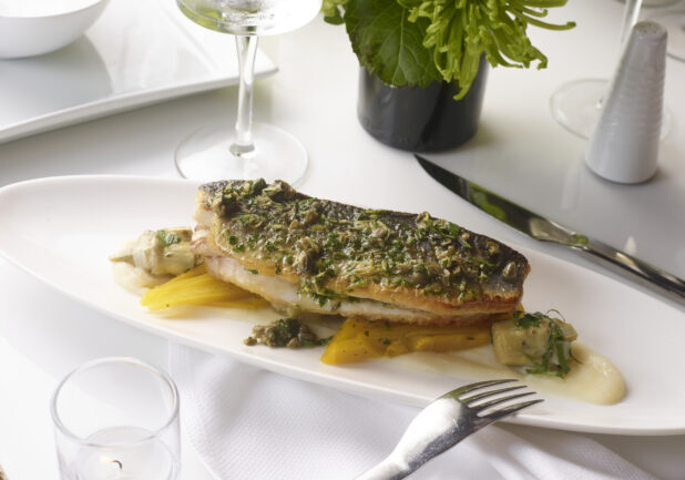 A White Ceramic Platter of Grilled Herb-Crusted Salmon or White Fish on a Bed of Roasted Yellow Carrots and White Cream Sauce on a White Table Cloth in a Restaurant Setting