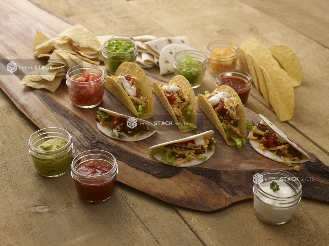 Taco platter with soft and hard tacos on a wooden platter with guacamole, salsa, sour cream, tomatoes, cheese and green onions in small mason jars with tortillas chips on the side