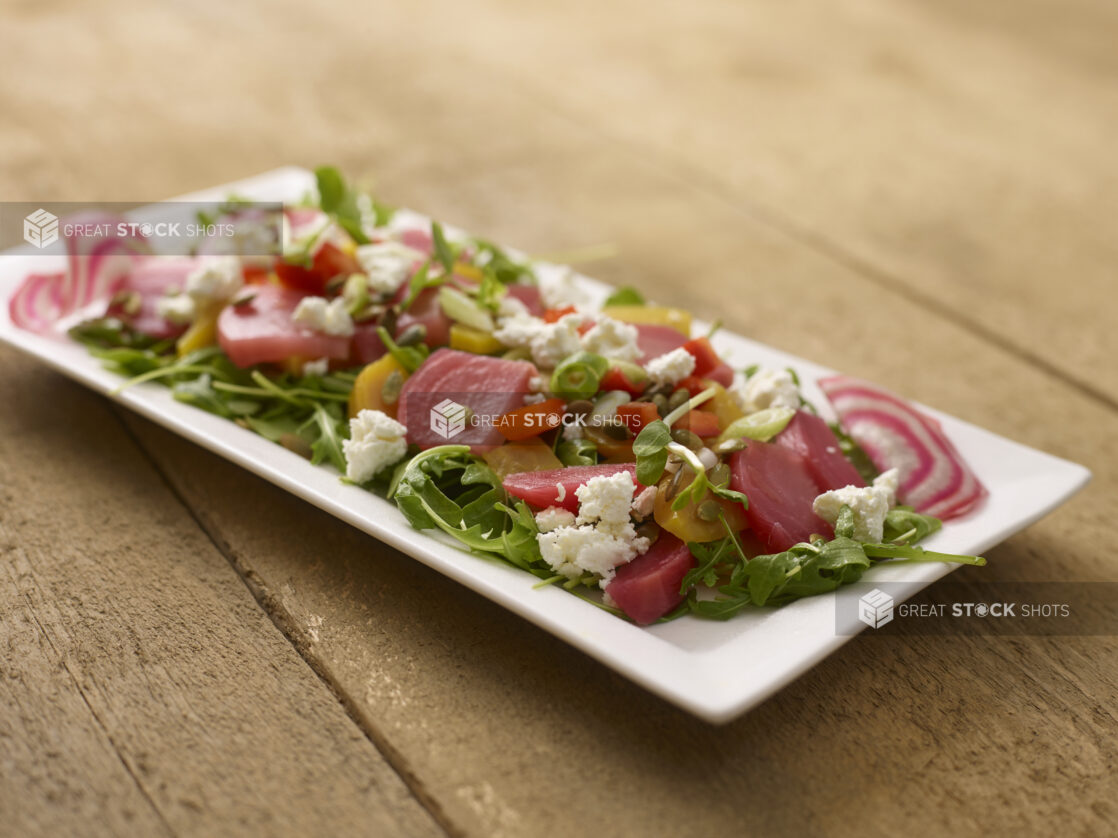 Mixed pickled beet salad on mixed greens with goats cheese on a white rectangular platter on a rustic wooden background