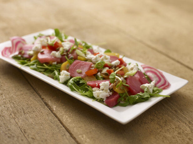Mixed pickled beet salad on mixed greens with goats cheese on a white rectangular platter on a rustic wooden background
