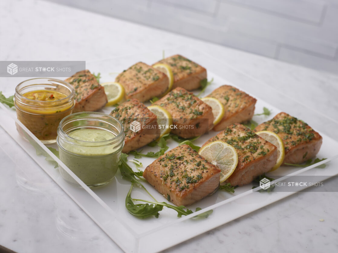 Pan seared salmon on a clear acrylic tray with lemon and arugula garnish with dill sauce and mango chutney in mason jars on a white marble background