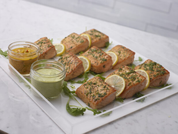 Pan seared salmon on a clear acrylic tray with lemon and arugula garnish with dill sauce and mango chutney in mason jars on a white marble background